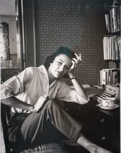 Imagen de Anne Sexton en su escritorio, apoyada sobre una mesa con libros y fumando