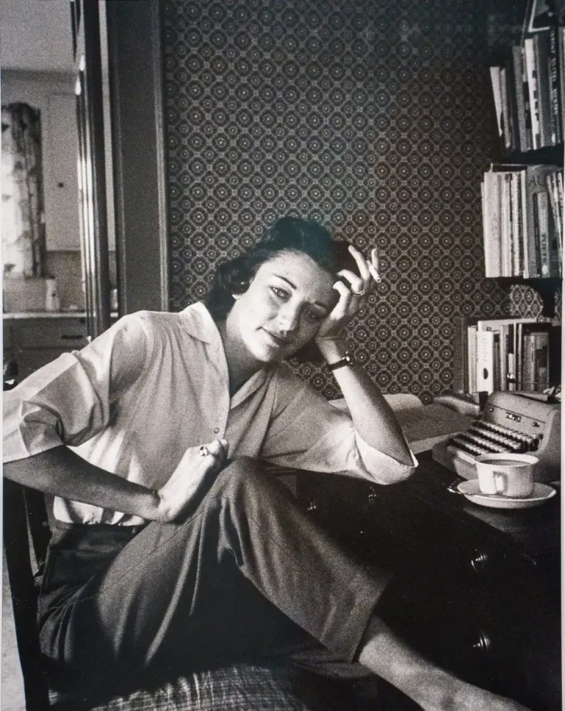 Imagen de Anne Sexton en su escritorio, apoyada sobre una mesa con libros y fumando