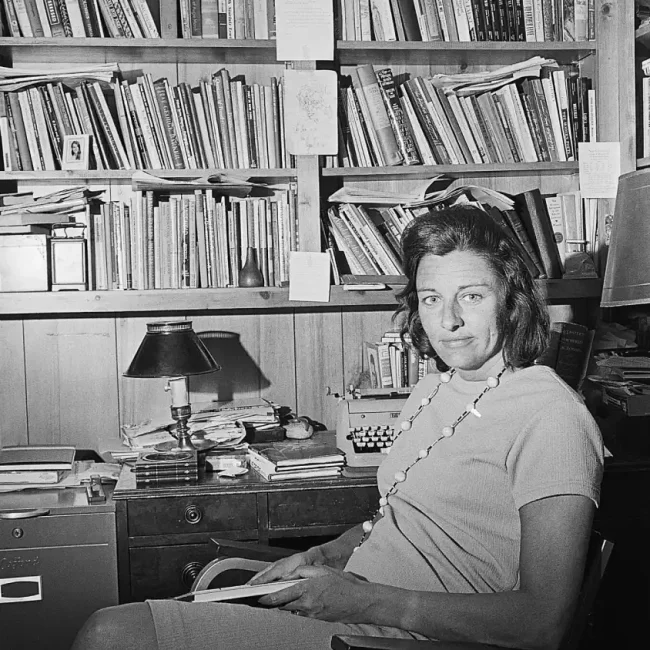 Anne Sexton en su despacho sentada y sonriente junto a una enorme biblioteca