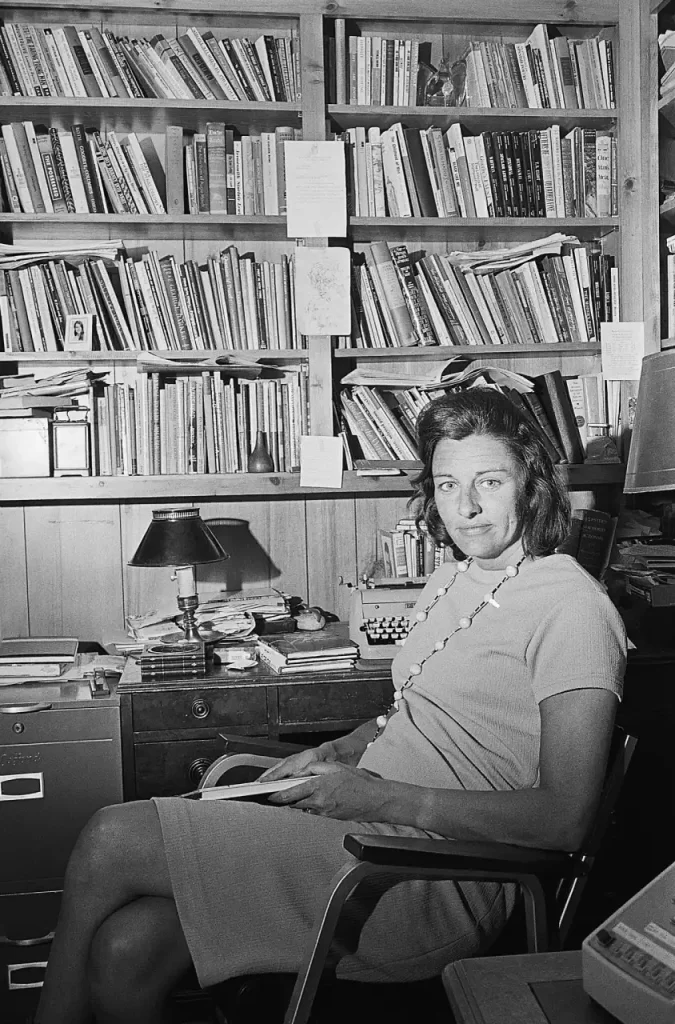 Anne Sexton en su despacho sentada y sonriente junto a una enorme biblioteca